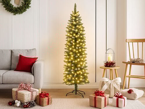 Egy világos, modern ünnepi jelenet, középen egy magas, karcsú, előre kivilágított karácsonyfával, meleg fehér fényekkel. Bal oldalon látható egy világosszürke kanapé karfája és háttámlája egy piros díszpárnával. Az előtérben egy világos szőnyegen ajándékcsomagok gyűjteménye pihen, piros és fehér szalagokkal, valamint tobozokkal díszítve. Jobbra egy fából készült szék és egy kis kerek fa oldalasztal áll, amelyen egy lámpás és gyertya található, teljessé téve a meghitt, ünnepi hangulatot.